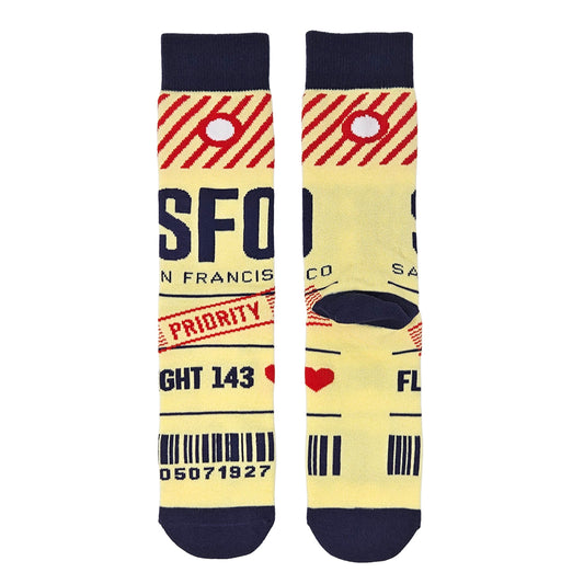 A pair of novelty crew socks with a vintage San Francisco airport (SFO) luggage tag design. The socks feature a pale yellow body with navy blue cuffs, heels, and toes, red diagonal stripes, a red priority stamp, and black printed text and barcodes.